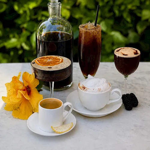 A sunny drink setup: a bottle of dark syrup, iced tea/coffee in glasses, espresso, lemon slice, blackberries, and yellow flowers.
