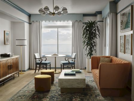 A bright living room with an ocean view, sheer curtains, a chandelier, two chairs, a couch, a coffee table, and a potted plant.