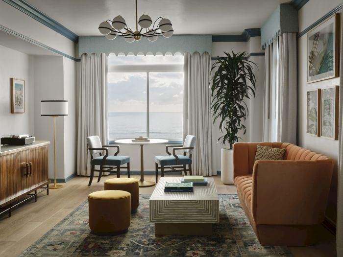 A bright living room with an ocean view, sheer curtains, a chandelier, two chairs, a couch, a coffee table, and a potted plant.