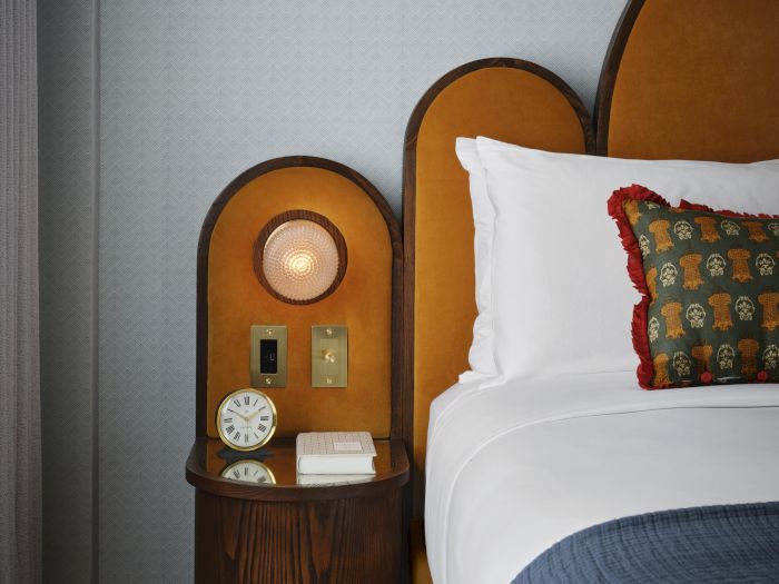 A cozy bedroom scene with a bedside lamp, alarm clock, and a bed dressed with white linens and a patterned decorative pillow.