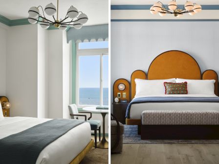 A bright hotel bedroom with two beds, a chandelier, and a view of the ocean through tall windows.