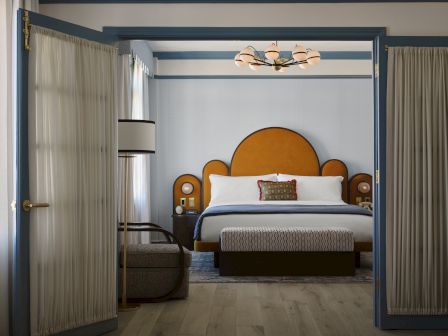 A cozy hotel bedroom viewed through open doors, with a large bed, bedside lamps, warm decor, and light curtains.