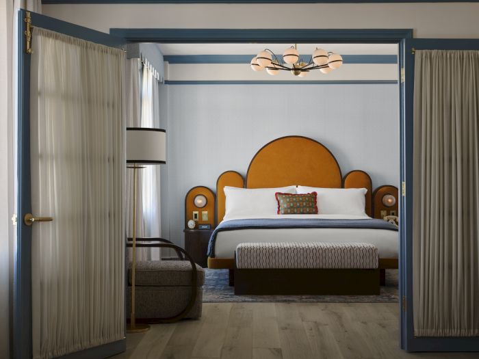 A cozy hotel bedroom viewed through open doors, with a large bed, bedside lamps, warm decor, and light curtains.
