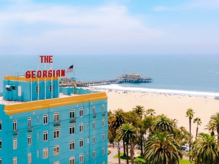 A beachfront hotel marked &ldquo;THE GEORGIAN&rdquo; overlooks a sandy beach, palms, and calm ocean under a bright sky.