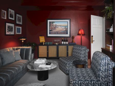 Cozy living room with red walls, framed &ldquo;Santa Monica&rdquo; art, a bar cabinet with bottles and lamps, plush couch, and patterned chairs.