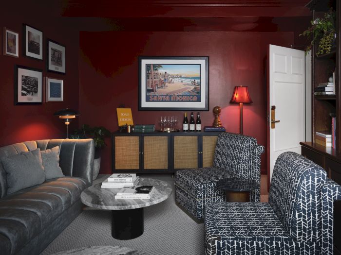 Cozy living room with red walls, framed &ldquo;Santa Monica&rdquo; art, a bar cabinet with bottles and lamps, plush couch, and patterned chairs.