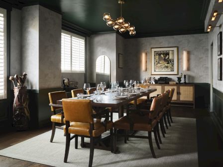 A stylish dining room set with a long table, leather chairs, wine glasses, wall art, and warm chandelier lighting.