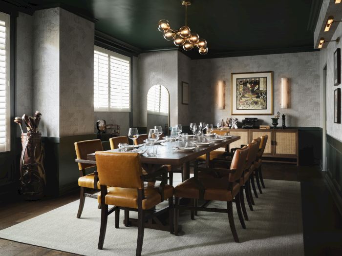 A stylish dining room set with a long table, leather chairs, wine glasses, wall art, and warm chandelier lighting.
