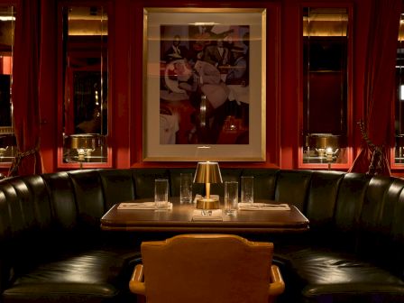 An intimate restaurant booth with red drapes, a framed mural, and a table set with glasses and a small lamp.