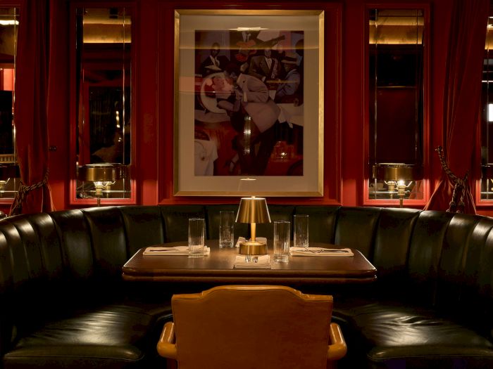 An intimate restaurant booth with red drapes, a framed mural, and a table set with glasses and a small lamp.