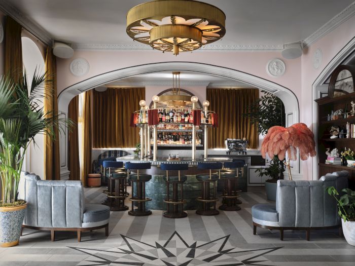 Stylish lounge bar interior with illuminated drink counter, stools, drapes, plants, chandeliers, and pink feather d&eacute;cor.