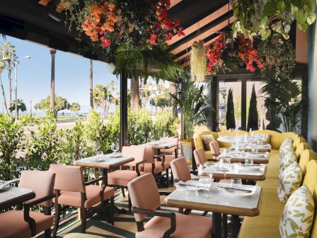 An outdoor dining patio with set tables, lush hanging plants and colorful flowers, and a sunny ocean view.