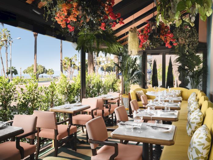 An outdoor dining patio with set tables, lush hanging plants and colorful flowers, and a sunny ocean view.