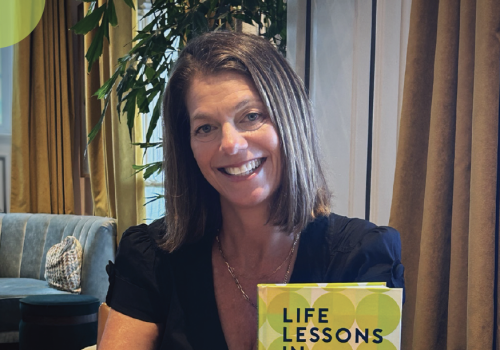 Smiling woman at home holding the book &ldquo;Life Lessons in Pickle-Ball,&rdquo; with several copies on the table.