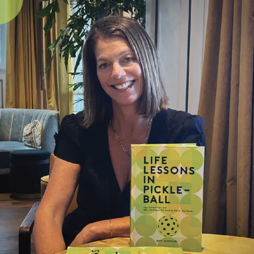 Smiling woman at home holding the book &ldquo;Life Lessons in Pickle-Ball,&rdquo; with several copies on the table.