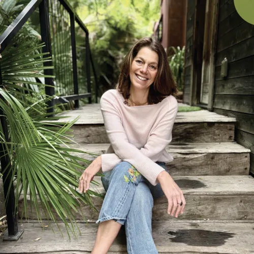 A smiling woman sits on outdoor wooden steps in a lush garden, wearing a pink sweater, jeans, and white sneakers.
