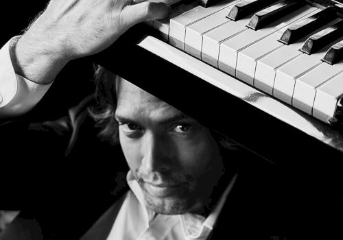 A man in a tuxedo plays a piano, his hand pressing the keys in a black-and-white shot.