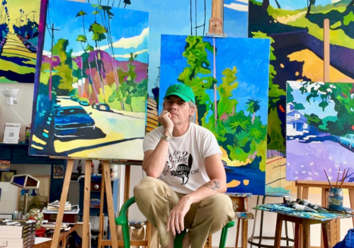 An artist in a green cap sits barefoot in a studio surrounded by colorful street-scape paintings on easels.