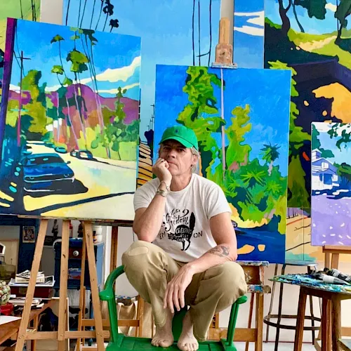 A man in an art studio sits on a stool in front of colorful canvases and paints, surrounded by brushes and easels.