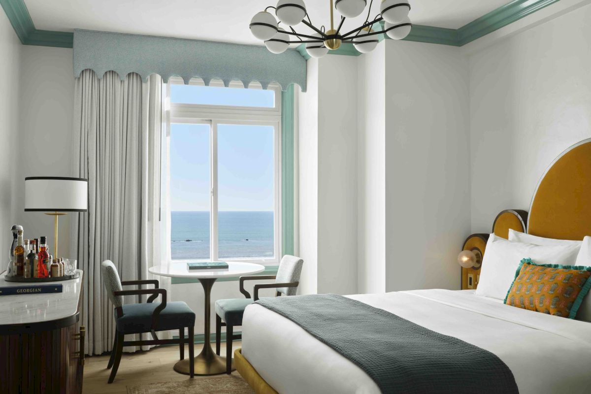 A bright seaside hotel room with a bed, chandelier, curtains, small table and lamp, and a view of the ocean through windows.