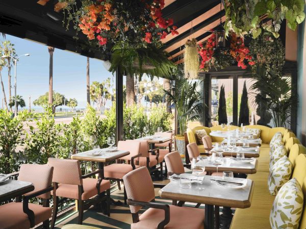 A sunny restaurant terrace with tables set for dining, lush hanging flowers and plants, and ocean views in the background.