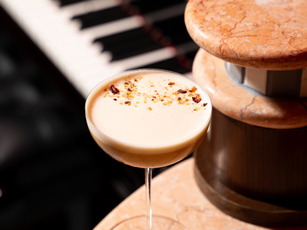 A creamy cocktail with a swirl and garnish sits on a marble bar top beside a shaker and blurred piano keys.