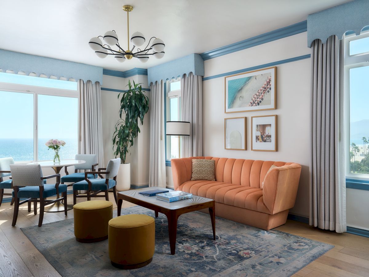 A bright living room with an orange sofa, coffee table, curtains, wall art, and large ocean-view windows, lit by a ceiling chandelier.