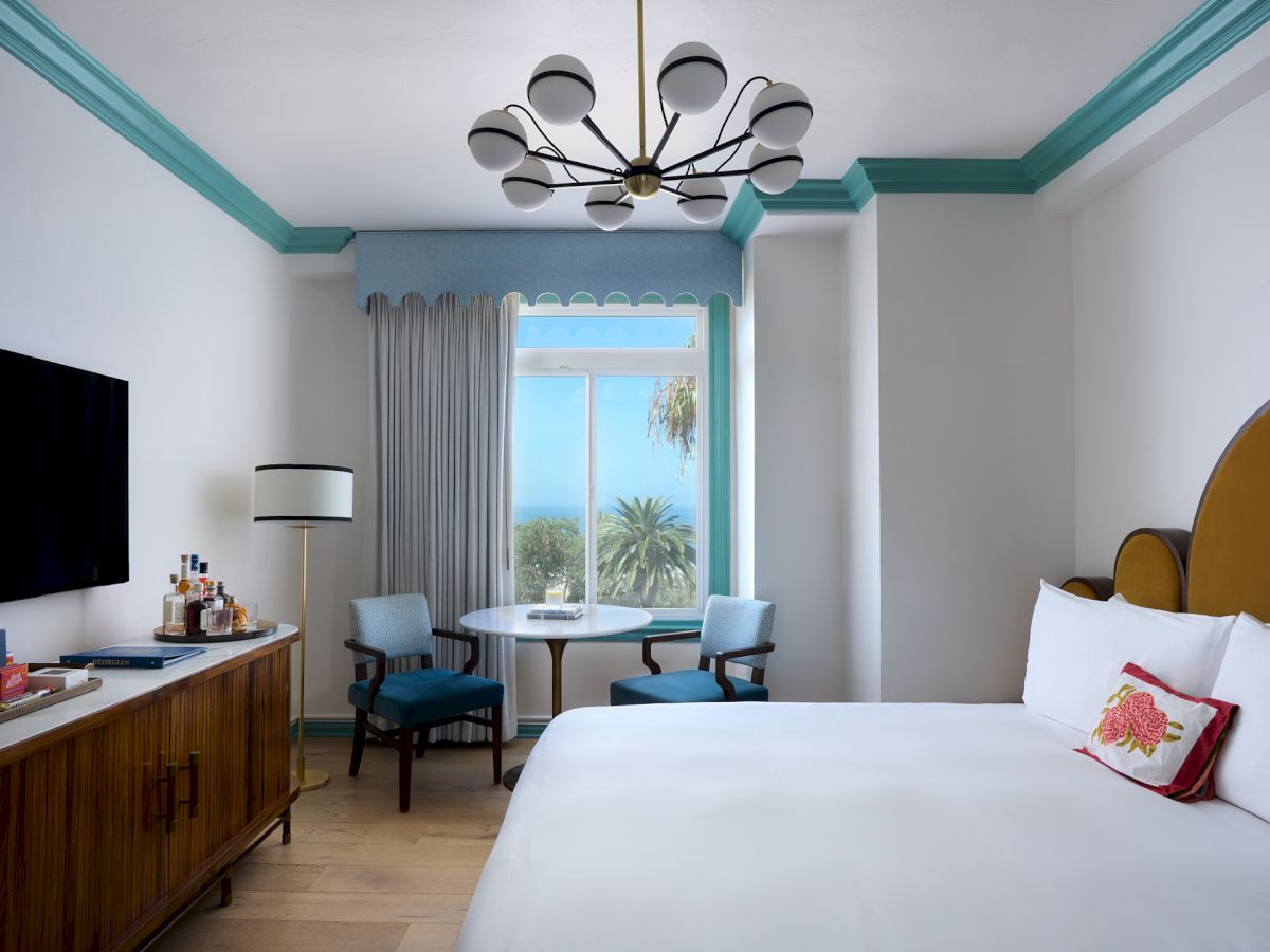A bright hotel room with a ceiling chandelier, bed, desk, small table and chairs, and teal-trim windows showing palm views.