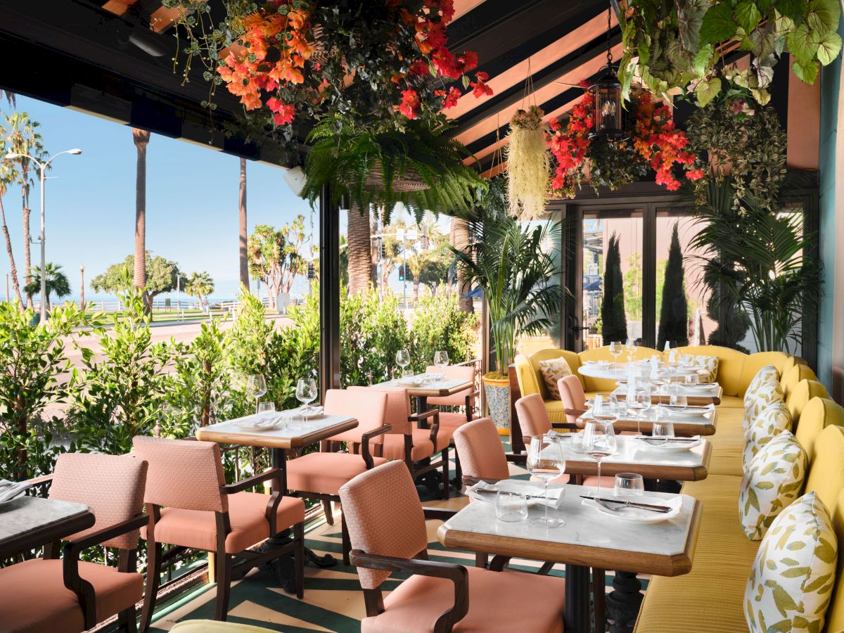 A sunny restaurant patio dining area with lush hanging plants, colorful flowers, and neatly set wooden tables and chairs outside.