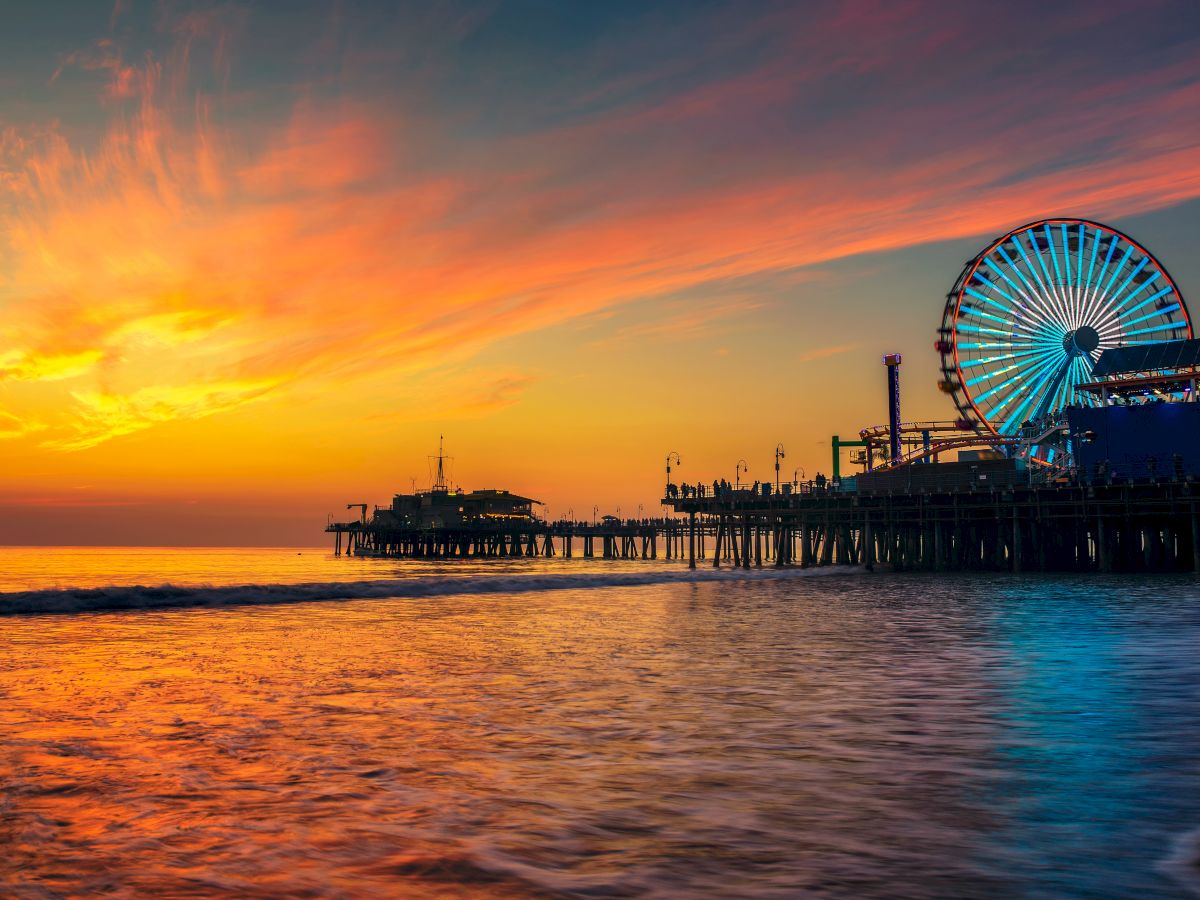 A Ferris wheel lights up beside a pier at sunset, with waves rolling onto the shore.