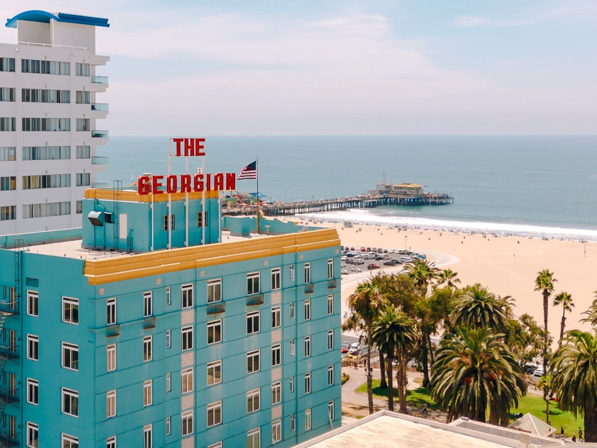 A seaside view of The George Inn hotel with palm trees, sandy beach, ocean, and a pier in the distance.