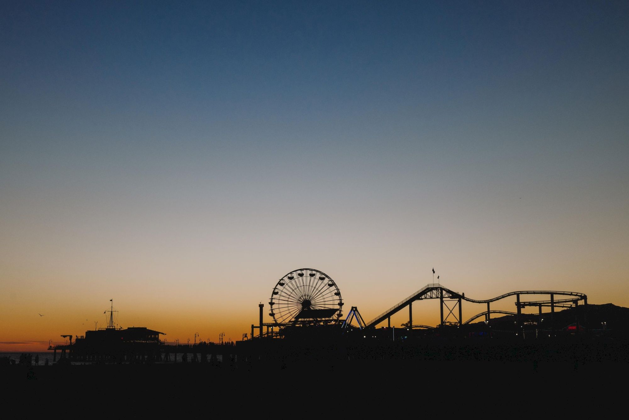 Silhouette of a Ferris wheel and roller coaster at a beach amusement pier during sunset.