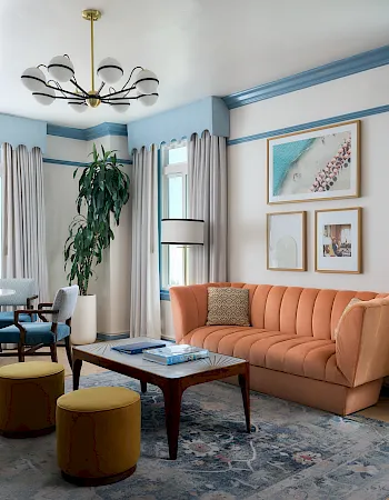 Bright living room with an ocean view, orange sofa, coffee table, dining chairs, curtains, framed art, and a chandelier.